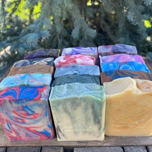 May include: A collection of rectangular bars of soap arranged in rows, featuring various colors including red, blue, green, brown, and purple with marbled patterns. Some bars have smooth surfaces, while others show textured tops and subtle swirls.