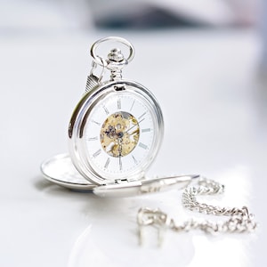May include: A silver pocket watch with an open face, revealing the intricate inner workings. The watch has a white face with black Roman numerals and delicate hands. A silver chain is attached. The watch is open, resting on a white surface.