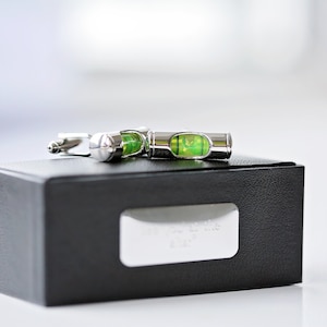 Può includere: Gemelli color argento con un design a livello in miniatura, con una bolla di liquido verde brillante. I gemelli sono appoggiati su una scatola nera con una targhetta argentata. La scatola ha una superficie strutturata.