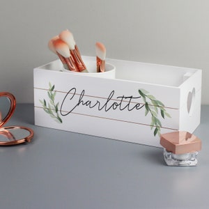 May include: White wooden box with a heart cutout and a gold leaf design. The box has the name "Charlotte" written on it in black cursive. The box is filled with makeup brushes and a small jar of cream.