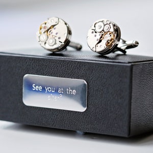 May include: A pair of silver cufflinks with exposed watch movement details, presented on a black box with a silver plaque. The plaque reads "See you at the altar?"