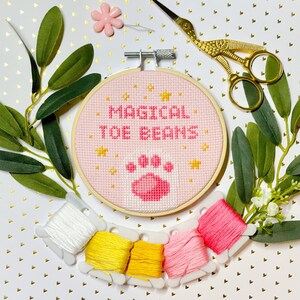 May include: A pink embroidery hoop with the text "Magical Toe Beans" and a pink paw print. The hoop is surrounded by white embroidery floss spools and green leaves.
