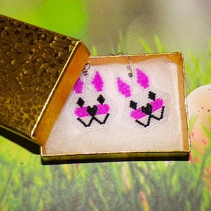 May include: A pair of beaded bunny earrings in a gold gift box. The earrings feature white beads with pink accents for the ears and nose, and black beads for the eyes and mouth. The box is open, revealing the earrings.