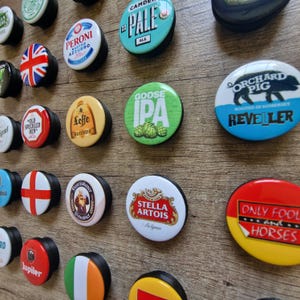 Peut inclure: Collection de bouchons de bouteille ronds avec des logos de marques de bière variés. Les bouchons présentent des logos tels que Peroni, Leffe, Stella Artois et Goose IPA. Les bouchons sont affichés sur une surface en bois.