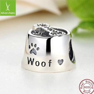 May include: Silver charm in the shape of a dog bowl with a bone on top. The bowl has a paw print and the word "Woof" with a heart.