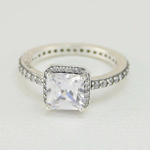 May include: A silver ring featuring a square-shaped clear cubic zirconia stone surrounded by smaller clear stones. The band is also set with smaller clear stones.