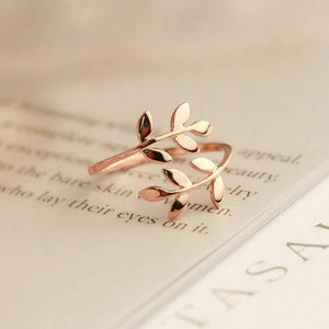 May include: A gold-toned ring featuring a delicate design of two intertwined branches with leaves. The ring is open and adjustable to fit different finger sizes.