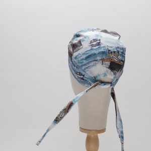May include: A white and blue patterned bandana with a design of ships and waves. The bandana has two long ties that can be tied at the back of the head.