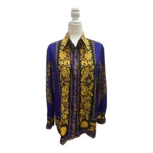 Könnte beinhalten: Ein langärmliges Button-Down-Hemd in leuchtendem Blau, verziert mit einem auffälligen goldenen und schwarzen Blumen- und Kettenmuster. Das Hemd wird auf einer Schaufensterpuppe präsentiert, die sein Design und seine Passform zeigt. Der Kragen und die Manschetten sind schwarz.
