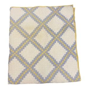 May include: A folded white quilt with a geometric pattern of blue and yellow diamonds. The quilt has a textured surface and a yellow border. The design is symmetrical and the colors are soft and inviting.
