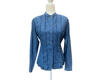 May include: A medium-wash blue denim shirt featuring a mandarin collar, long sleeves, and a front button-down closure. The shirt is designed with vertical pleated detailing running down the front panels, creating a structured and textured appearance.