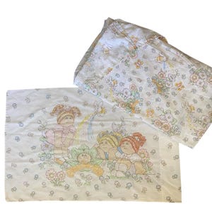 Pode incluir: Conjunto de lençóis vintage Cabbage Patch Kids de solteiro. Os lençóis apresentam um padrão repetido de Cabbage Patch Kids, flores e borboletas em cores pastel sobre um fundo branco. O lençol de cima tem uma imagem maior das bonecas.