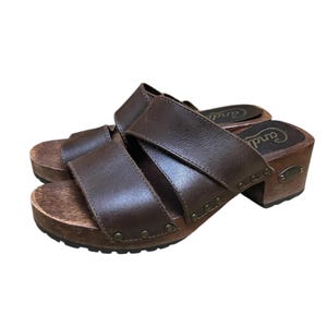Könnte beinhalten: Ein Paar braune Lederclogs mit Holzsohlen. Die Clogs haben zwei Riemen über dem Oberteil und einen kleinen Absatz. Das Wort "Candy" ist auf der Seite der Clogs sichtbar.