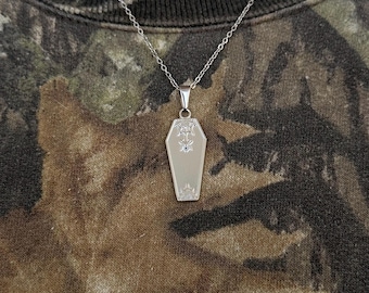 May include: A silver chain necklace with a silver coffin pendant. The coffin has a detailed design and a small diamond-like stone in the center.