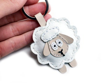 May include: A white leather sheep keychain with black eyes and a beige face. The sheep has a silver ring attached to its back.