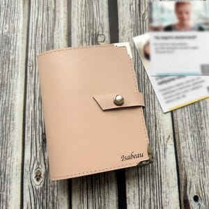 May include: A pale pink leather wallet with a snap closure and silver corner protectors. The name "Isabeau" is engraved on the front. Two business cards are in the background on a wooden surface.