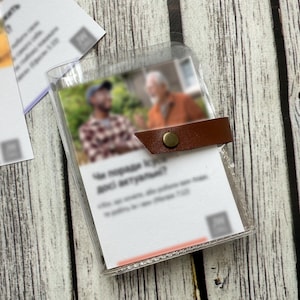 May include: A clear plastic card holder with a brown leather strap and a brass button closure. The holder contains a business card with blurred text and images. Additional cards are visible in the background.