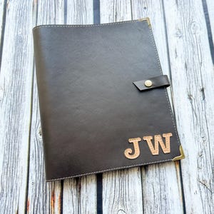Puede incluir: Un diario de cuero marrón oscuro con cierre de botón a presión y protectores de esquina dorados. Las iniciales "JW" se muestran en un color marrón más claro en la portada. El diario está sobre una superficie de madera clara.
