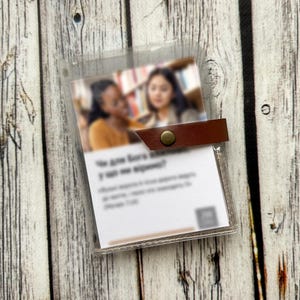 May include: A transparent plastic card holder with a brown leather strap and a brass stud fastener. Inside is a printed card with text and a blurred image of two women. The background is a wooden surface.