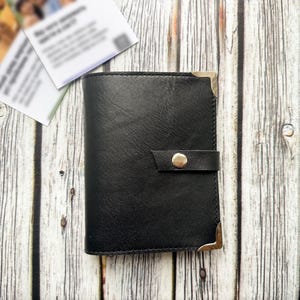 May include: A black leather wallet with a snap closure and metal corner protectors. The wallet is closed and has a simple, classic design. Two white cards are in the background.