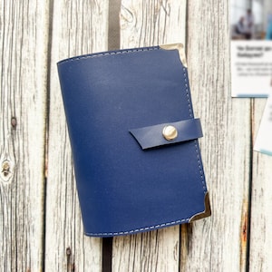 May include: A navy blue leather wallet with a gold snap closure and corner accents. The wallet is closed with a strap and button. The stitching is white. The wallet is on a wooden surface.