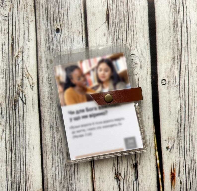 May include: A clear plastic card holder with a brown leather strap and a brass snap closure. The holder contains a printed card with text and a blurred image of two women. The background is a wooden surface.