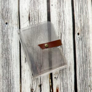 May include: A clear, transparent plastic wallet with a brown leather strap closure and a gold-colored snap. The wallet is designed to hold cards or other small items. The background is a light-colored wooden surface.