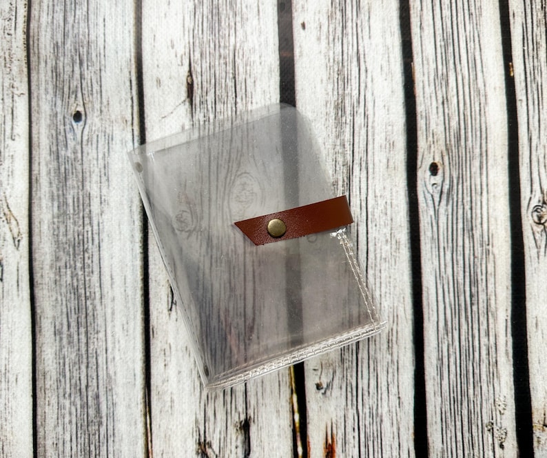 May include: A clear, transparent plastic wallet with a brown leather strap closure and a gold-colored snap. The wallet is designed to hold cards or other small items. The background is a light-colored wooden surface.