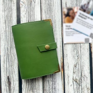 May include: A green leather journal with a brass button closure and corner accents. The journal is closed and sits on a wooden surface. A business card is in the background.