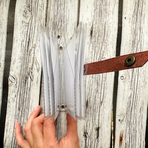 May include: A clear plastic card holder with multiple pockets, held open to display the contents. A brown leather strap with a metal snap closure is attached to the side. The background is a light-colored wooden surface.
