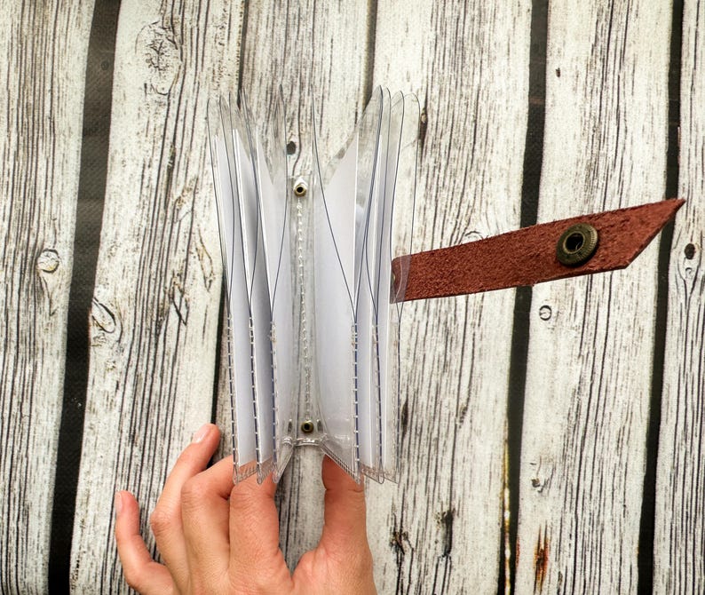 May include: A clear plastic card holder with multiple pockets, held open to display the contents. A brown leather strap with a metal snap closure is attached to the side. The background is a light-colored wooden surface.