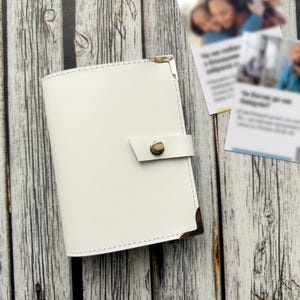 May include: A white leather journal with a snap closure and metal corner protectors. The journal is on a wooden surface, with two blurred cards in the background. The journal's design is simple and elegant.