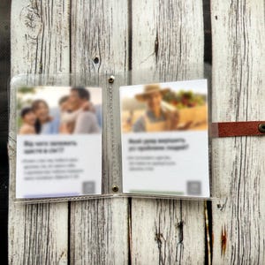 May include: A clear plastic photo holder with two inserts, secured by metal rivets. A brown leather strap with a metal snap closure is attached to the side. The background is a light-colored wooden surface.