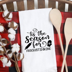 May include: A white kitchen towel with the text "tis the season for ProcrastiBaking" in black script, a rolling pin, and a mitten graphic. The towel is in a wooden basket with a red and black plaid cloth, cotton sprigs, and wooden spoons.