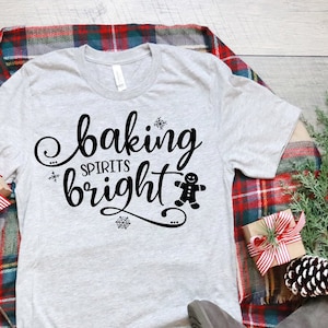 May include: A grey t-shirt with the text "baking spirits bright" and a gingerbread man graphic. The shirt is laid out on a wooden surface with a plaid blanket, blue jeans, and brown boots.