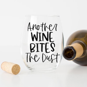 May include: A clear glass wine glass with the text "Another Wine Bites The Dust" printed on it. A cork and a bottle of wine are also in the image.