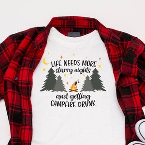 May include: White t-shirt with a camping-themed graphic that says "Life needs more starry nights and getting campfire drunk". The shirt is laid out on a white surface with a red and black plaid shirt and white sneakers.