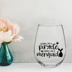 May include: A clear glass wine glass with a black vinyl decal that reads "Drink like a pirate, dance like a mermaid". The decal includes a skull and crossbones and a mermaid tail.