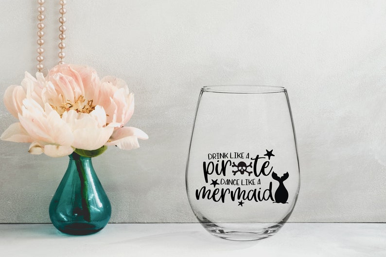 May include: A clear glass wine glass with a black vinyl decal that reads "Drink like a pirate, dance like a mermaid". The decal includes a skull and crossbones and a mermaid tail.