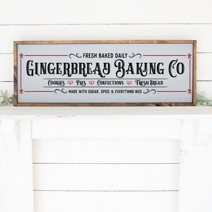Pu&ograve; includere: Un cartello rettangolare con cornice in legno e sfondo grigio chiaro che presenta il testo nero GINGERBREAD BAKING CO, FRESH BAKED DAILY, COOKIES, PIES, CONFECTIONS, FRESH BREAD e MADE WITH SUGAR, SPICE & EVERYTHING NICE.