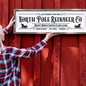 Pu&ograve; includere: Un cartello rettangolare in legno con sfondo bianco e cornice marrone scuro. Il testo recita: Old Fashioned Sleigh Rides, North Pole Reindeer Co, Right Down Santa Claus Lane, Warm Blanket, Hot Cocoa, Memories, con due icone di slitta nere.