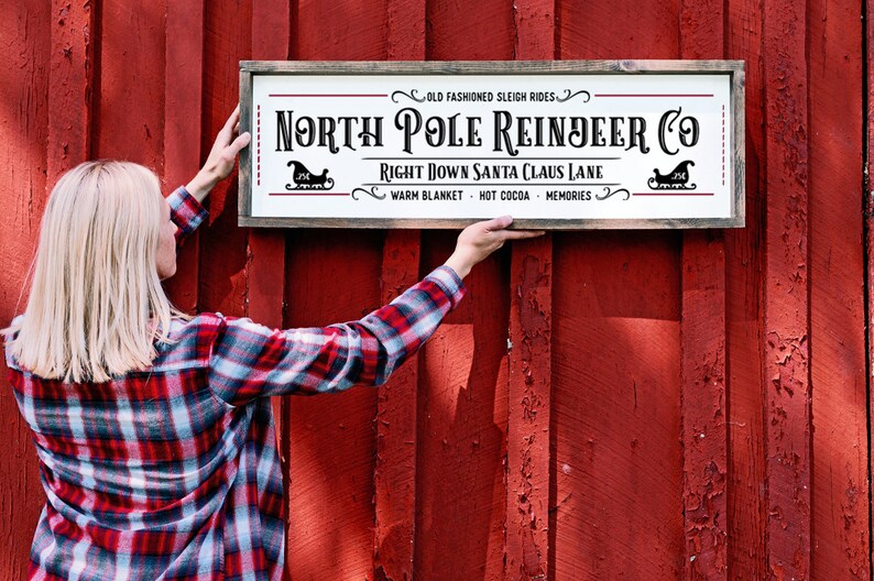 Pu&ograve; includere: Un cartello rettangolare in legno con sfondo bianco e cornice marrone scuro. Il testo recita: Old Fashioned Sleigh Rides, North Pole Reindeer Co, Right Down Santa Claus Lane, Warm Blanket, Hot Cocoa, Memories, con due icone di slitta nere.
