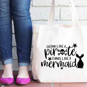 May include: A white canvas tote bag with black text that reads "Drink like a pirate, dance like a mermaid". The text is surrounded by stars and a skull and crossbones. A mermaid tail is also visible on the bag.