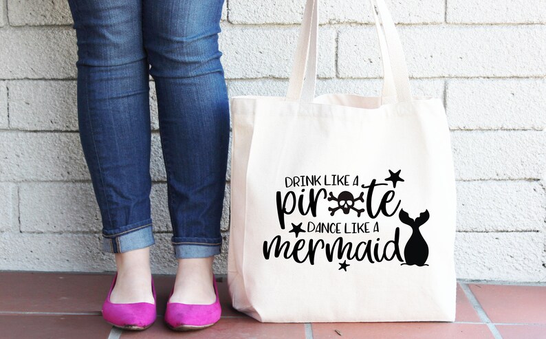 May include: A white canvas tote bag with black text that reads "Drink like a pirate, dance like a mermaid". The text is surrounded by stars and a skull and crossbones. A mermaid tail is also visible on the bag.