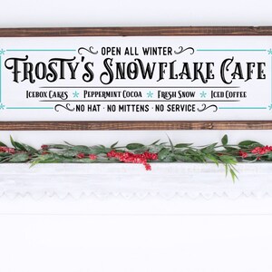 Pu&ograve; includere: Un'insegna rettangolare in legno con cornice marrone scuro e sfondo bianco con testo nero che recita Frosty's Snowflake Cafe, Open All Winter, Icebox Cakes, Peppermint Cocoa, Fresh Snow, Iced Coffee e No Hat No Mittens No Service.
