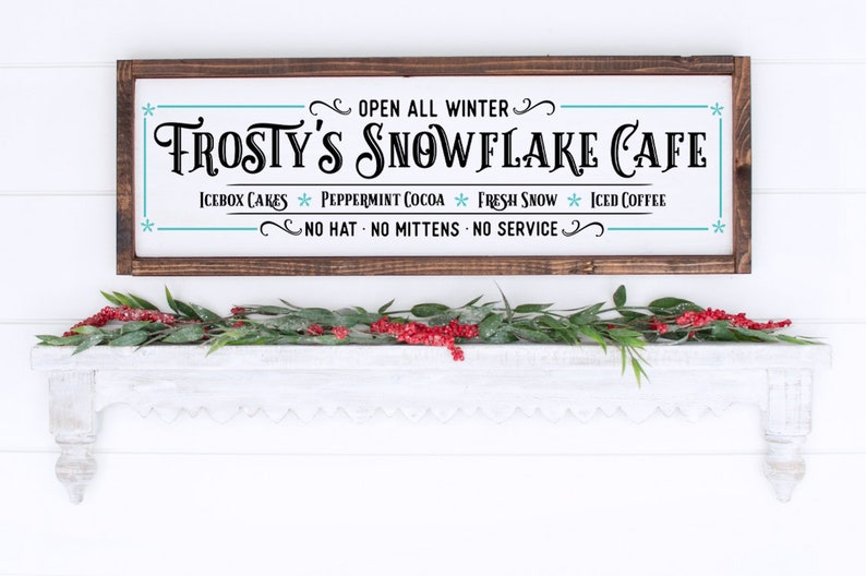 Pu&ograve; includere: Un'insegna rettangolare in legno con cornice marrone scuro e sfondo bianco con testo nero che recita Frosty's Snowflake Cafe, Open All Winter, Icebox Cakes, Peppermint Cocoa, Fresh Snow, Iced Coffee e No Hat No Mittens No Service.