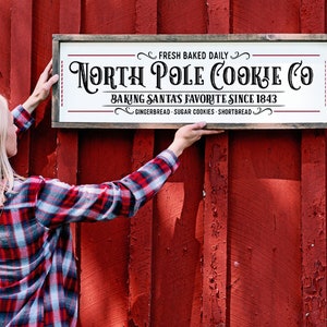 Pu&ograve; includere: Un cartello rettangolare in legno con sfondo bianco e cornice scura. Il testo recita: FRESH BAKED DAILY, NORTH POLE COOKIE CO, BAKING SANTAS FAVORITE SINCE 1843, GINGERBREAD - SUGAR COOKIES - SHORTBREAD. Il cartello presenta decorazioni ornamentali.
