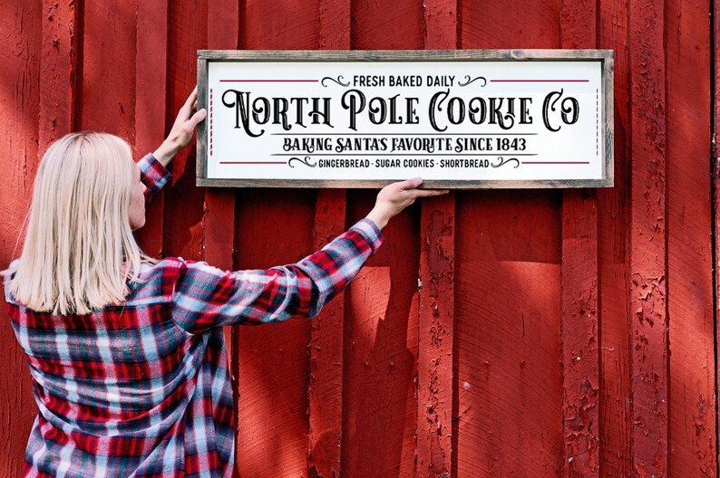 Pu&ograve; includere: Un cartello rettangolare in legno con sfondo bianco e cornice scura. Il testo recita: FRESH BAKED DAILY, NORTH POLE COOKIE CO, BAKING SANTAS FAVORITE SINCE 1843, GINGERBREAD - SUGAR COOKIES - SHORTBREAD. Il cartello presenta decorazioni ornamentali.
