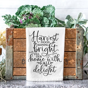 Może przedstawiać: Biały ręcznik kuchenny z czarnym tekstem, kt&oacute;ry brzmi "Harvest moon shining bright fill our home with fall delight". Ręcznik leży na drewnianej skrzyni z zielonymi roślinami po obu stronach.