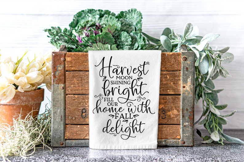 Może przedstawiać: Biały ręcznik kuchenny z czarnym tekstem, kt&oacute;ry brzmi "Harvest moon shining bright fill our home with fall delight". Ręcznik leży na drewnianej skrzyni z zielonymi roślinami po obu stronach.
